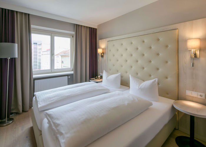 Vista della camera doppia Basic dell'Hotel Sailer Innsbruck con un letto matrimoniale, una lampada da terra, pavimento in parquet e una grande finestra.