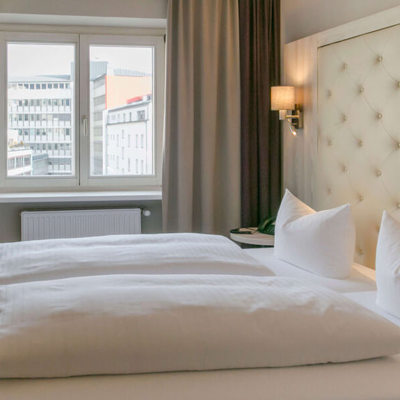 Blick in das Doppelzimmer Basic im Hotel Sailer Innsbruck mit einem Doppelbett, einer Stehlampe, Parkettboden und einem großen Fenster