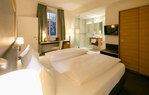 A comfortable bed stands in the middle of a suite. The room has a bathroom with glass doors and a door to the second room.