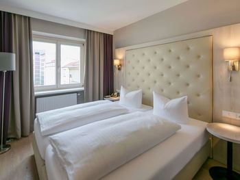 Vista della camera doppia Basic dell'Hotel Sailer Innsbruck con un letto matrimoniale, una lampada da terra, pavimento in parquet e una grande finestra.