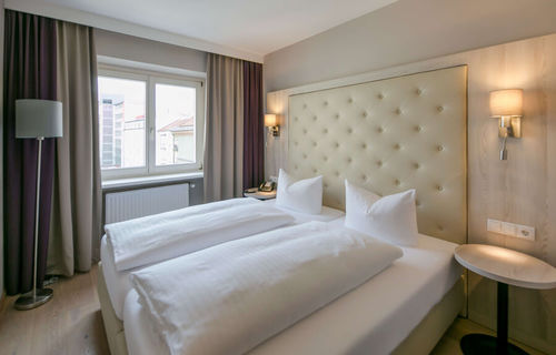 Vista della camera doppia Basic dell'Hotel Sailer Innsbruck con un letto matrimoniale, una lampada da terra, pavimento in parquet e una grande finestra.