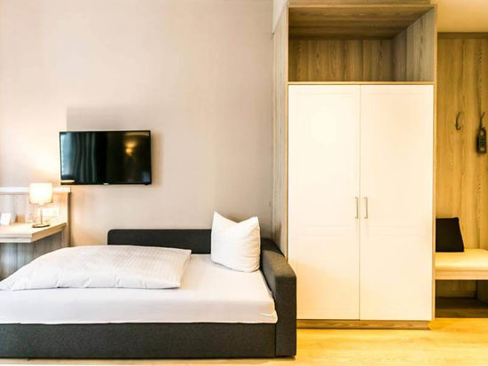 Ein Couchbett steht neben einem Schrank und einer feinen Sitzgelegenheit. Über dem Bett ist ein Flachbildschirm-Fernseher.