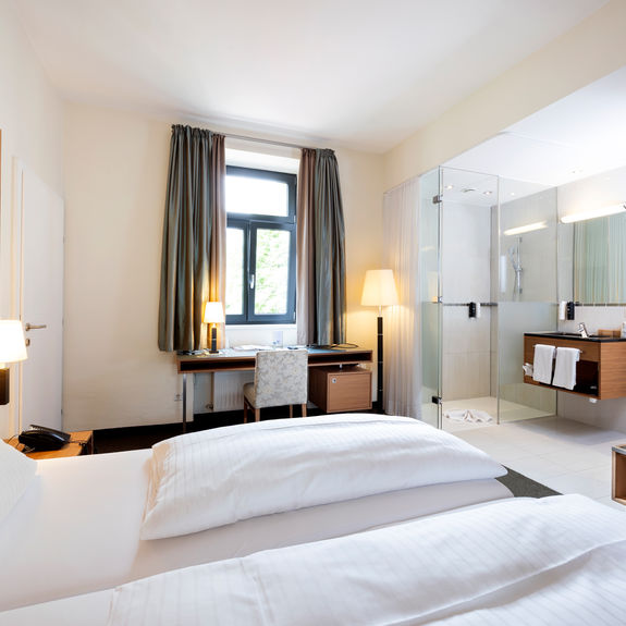 Ein gemütliches Bett steht der Mitte einer Suite. Das Zimmer hat ein Badezimmer mit Glaswänden und einen Tür ins zweite Zimmer.