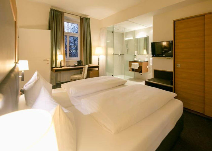 A comfortable bed stands in the middle of a suite. The room has a bathroom with glass doors and a door to the second room.