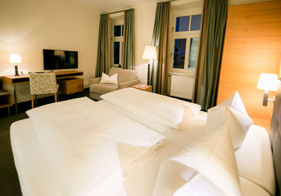 Vista nella camera business dell'Hotel Sailer con un letto matrimoniale, un comodo divano, una lampada da terra e una TV a schermo piatto