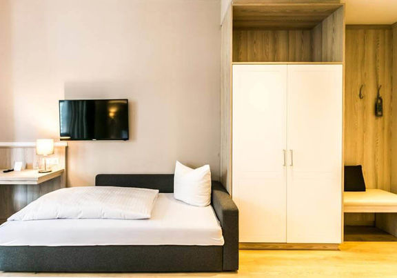 Ein Couchbett steht neben einem Schrank und einer feinen Sitzgelegenheit. Über dem Bett ist ein Flachbildschirm-Fernseher.