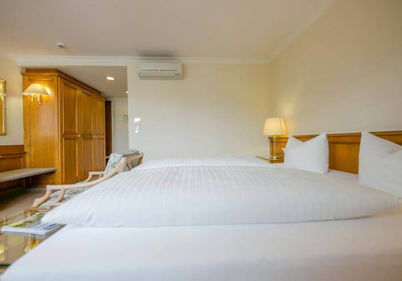 Weiche Doppelbetten befinden sich in einen geräumigen Hotelzimmer. Im Zimmer stehet ein großer Kleiderschrank und eine Sitzgelegenheit.