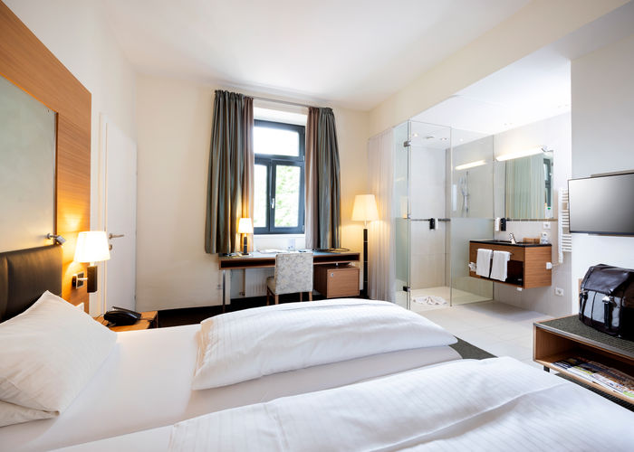 Ein gemütliches Bett steht der Mitte einer Suite. Das Zimmer hat ein Badezimmer mit Glaswänden und einen Tür ins zweite Zimmer.