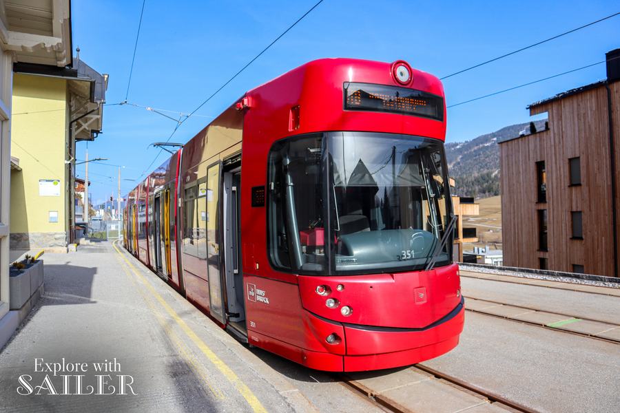 Eine rote moderne Straßenbahn der Linie STB steht an einer Haltestelle in Fulpmes vor alpiner Kulisse.