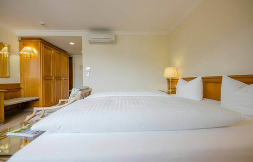 Weiche Doppelbetten befinden sich in einen geräumigen Hotelzimmer. Im Zimmer stehet ein großer Kleiderschrank und eine Sitzgelegenheit.