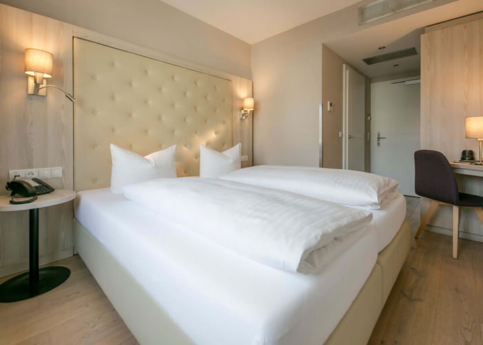 Vista nella camera doppia Basic dell'Hotel Sailer con un grande letto matrimoniale, lenzuola bianche, una scrivania e una sedia