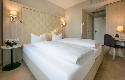 Vista nella camera doppia Basic dell'Hotel Sailer con un grande letto matrimoniale, lenzuola bianche, una scrivania e una sedia