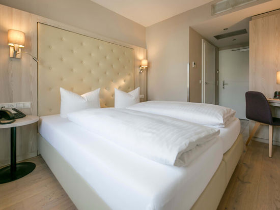 Vista nella camera doppia Basic dell'Hotel Sailer con un grande letto matrimoniale, lenzuola bianche, una scrivania e una sedia