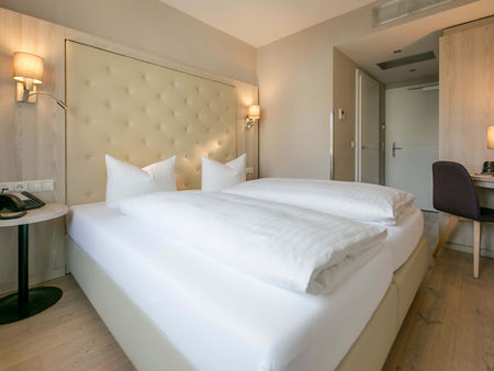 Vista nella camera doppia Basic dell'Hotel Sailer con un grande letto matrimoniale, lenzuola bianche, una scrivania e una sedia