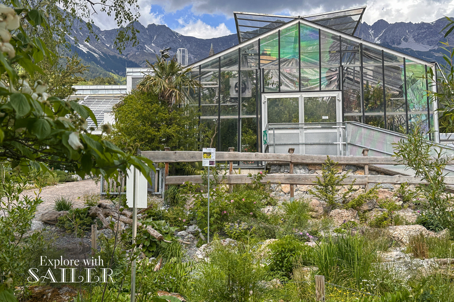 Glasgewächshaus umgeben von Pflanzen und alpiner Landschaft mit Bergen im Hintergrund.
