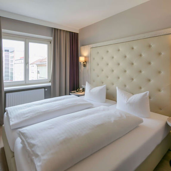 Blick in das Doppelzimmer Basic im Hotel Sailer Innsbruck mit einem Doppelbett, einer Stehlampe, Parkettboden und einem großen Fenster