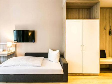 Ein Couchbett steht neben einem Schrank und einer feinen Sitzgelegenheit. Über dem Bett ist ein Flachbildschirm-Fernseher.