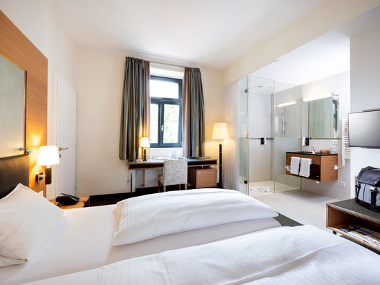 Ein gemütliches Bett steht der Mitte einer Suite. Das Zimmer hat ein Badezimmer mit Glaswänden und einen Tür ins zweite Zimmer.