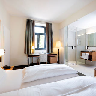 Ein gemütliches Bett steht der Mitte einer Suite. Das Zimmer hat ein Badezimmer mit Glaswänden und einen Tür ins zweite Zimmer.