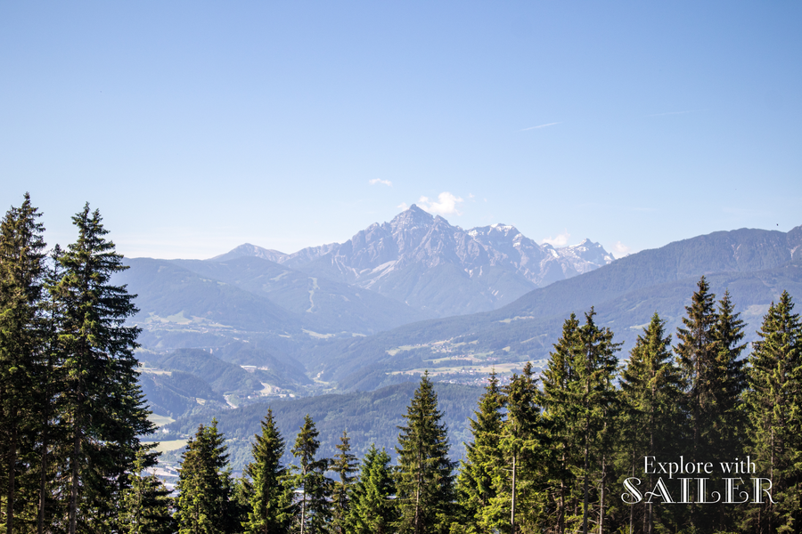Weitläufiger Blick auf die Tiroler Bergwelt und das Tal mit einem Nadelwald im Vordergrund unter klarem Himmel.