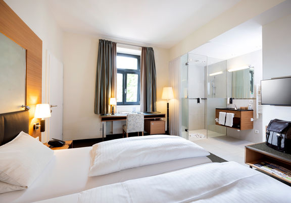 Ein gemütliches Bett steht der Mitte einer Suite. Das Zimmer hat ein Badezimmer mit Glaswänden und einen Tür ins zweite Zimmer.