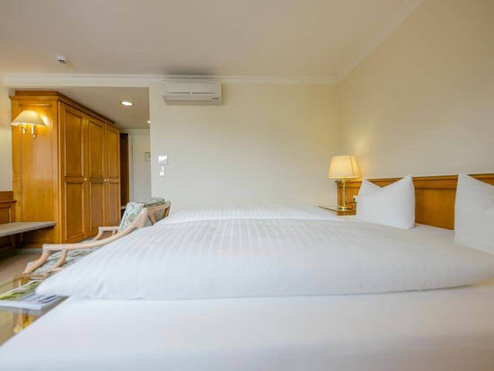 Weiche Doppelbetten befinden sich in einen geräumigen Hotelzimmer. Im Zimmer stehet ein großer Kleiderschrank und eine Sitzgelegenheit.