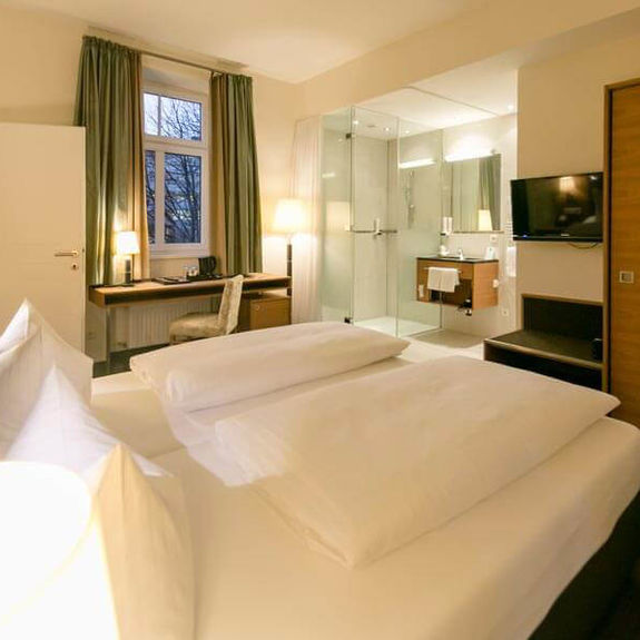 A comfortable bed stands in the middle of a suite. The room has a bathroom with glass doors and a door to the second room.