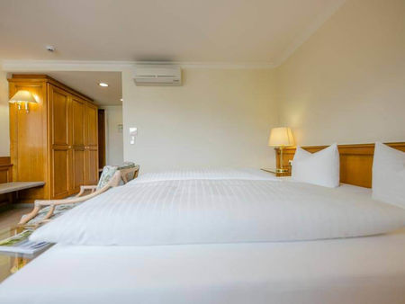 Weiche Doppelbetten befinden sich in einen geräumigen Hotelzimmer. Im Zimmer stehet ein großer Kleiderschrank und eine Sitzgelegenheit.