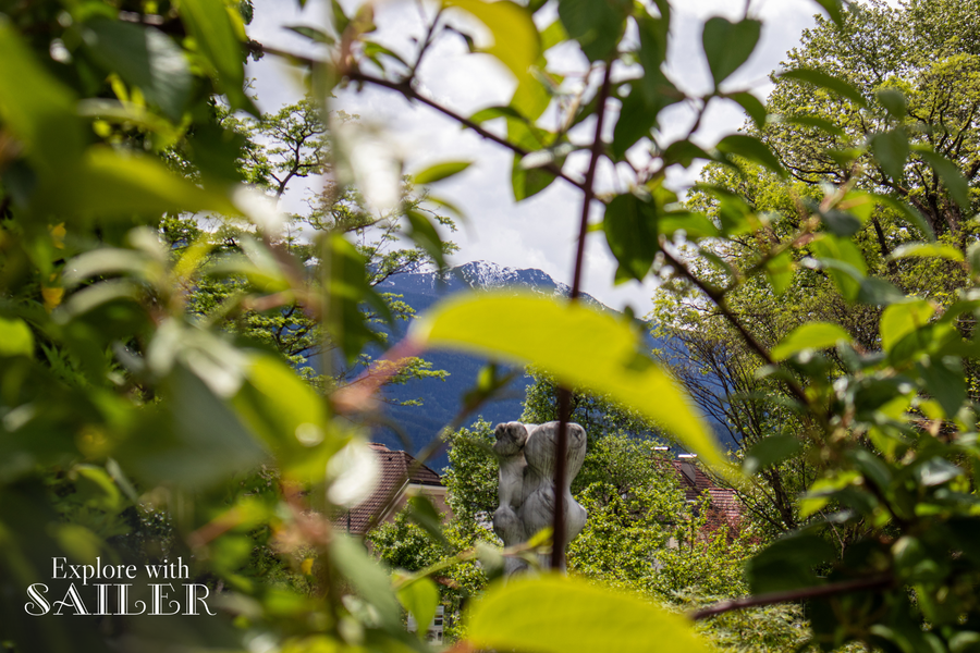 Grüne Blätter und Äste rahmen den Blick auf alpine Berge und eine Skulptur im Botanischen Garten bei sonnigem Wetter ein.