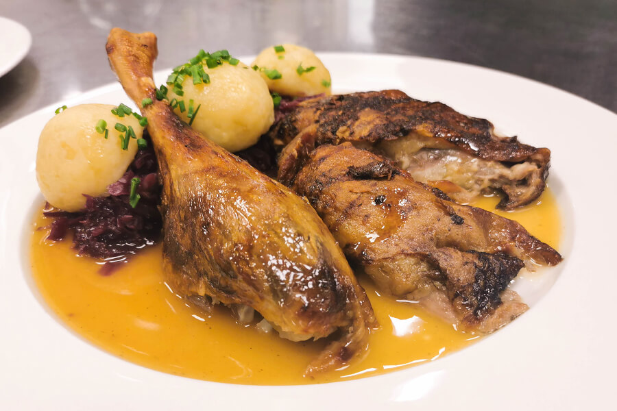Ein gebratener Gänse-Schenkel und zwei gebratene Gänse-Brüste liegen neben Rotkraut und drei Kartoffelknödeln in einer braunen Sauce auf einem weißen Teller