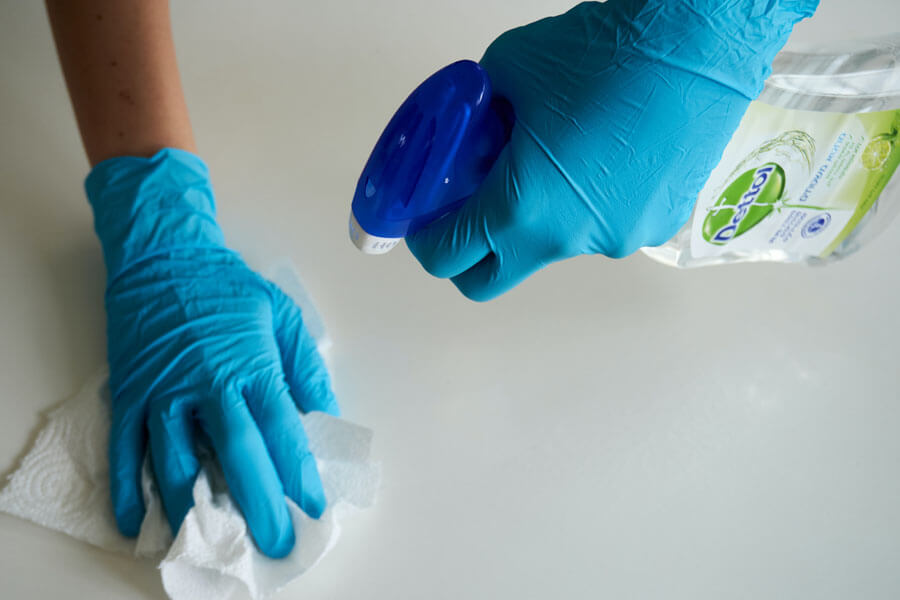 A woman with cleaning gloves cleaning a white surface with paper and a disinfectant spray