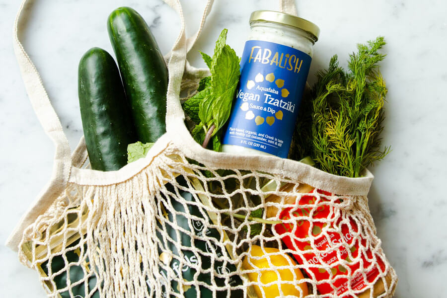 Zucchini, Tzatziki, Zitronen, eine Dose mit gehackten Tomaten und mehr liegen in einem Einkaufsnetz auf einer weiß-grauen Marmorplatte