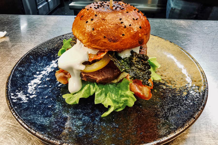 Ein Burger mit Salatblättern, einer gebratenen Scheibe Hackfleisch, roten und gelben Tomaten, einem etwas angebackenen Brot und einem weißen Dressing liegt auf einem dunkelgrauen Teller