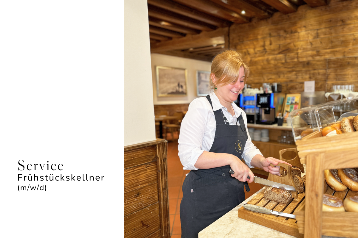 Ein körniges Brot mit einem weißen Aufstrich, einer Scheibe Käse und drei Scheiben Gurke liegt auf einem weißen Untergrund