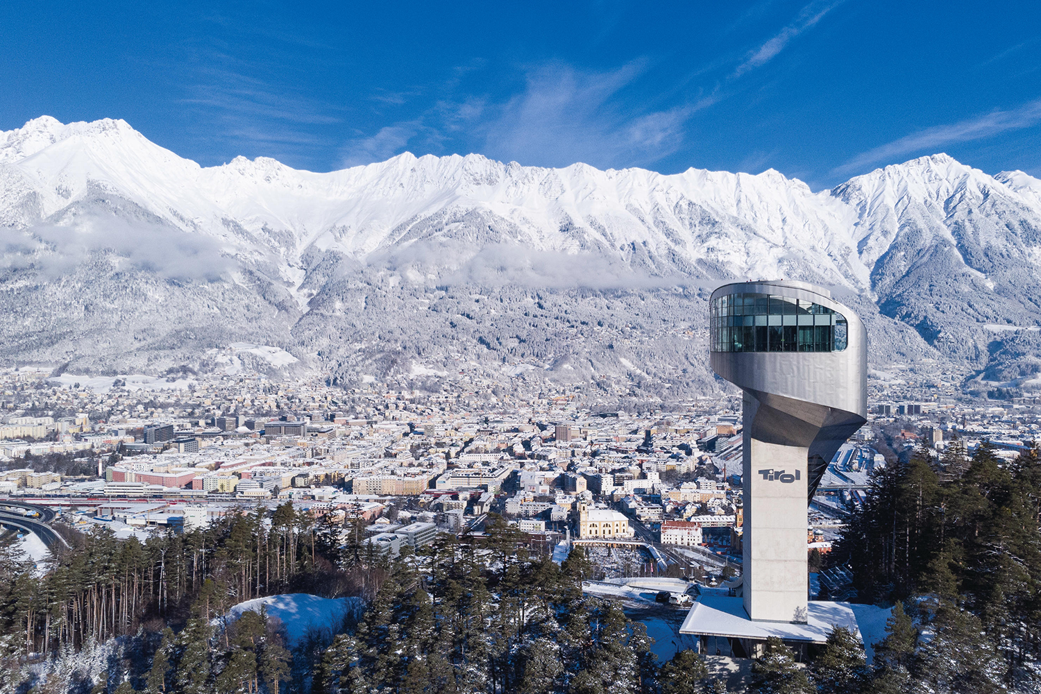 Snowy mountain landscape with modern observation tower and city view in the foreground.