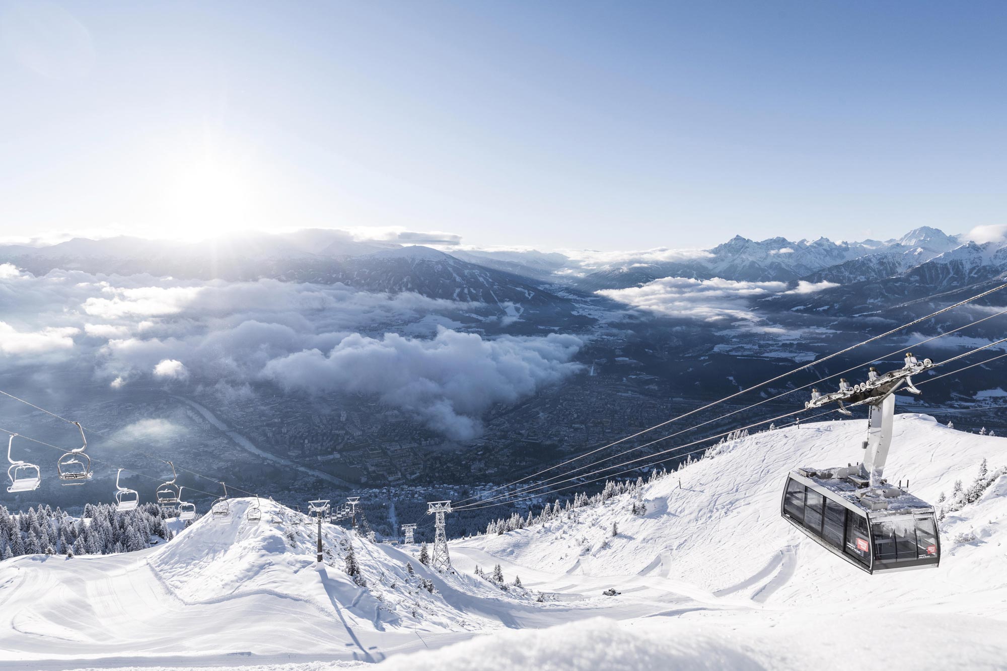Seilbahn über schneebedeckten Bergen mit strahlendem Sonnenschein und wolkenverhangenem Tal.