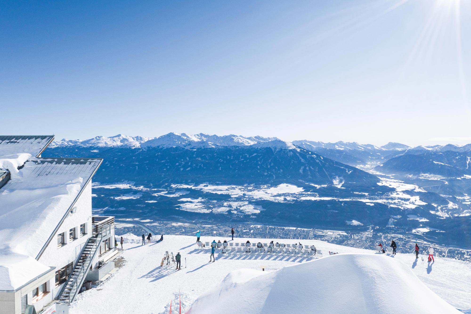 Schneebedeckte Alpen mit Sonnenschein, Menschen genießen die Aussicht von einer Terrasse.