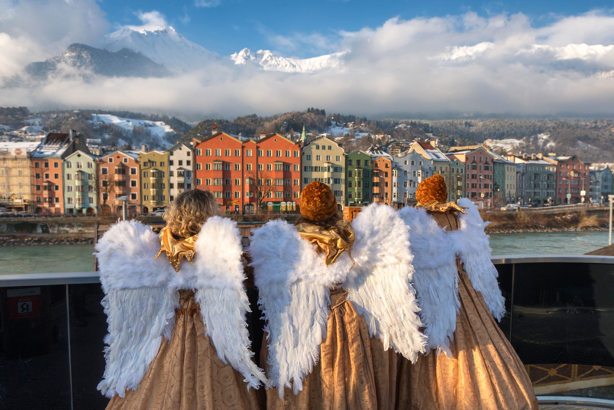Drei kostümierte Engel blicken auf bunte Häuserzeile vor schneebedeckten Alpen.