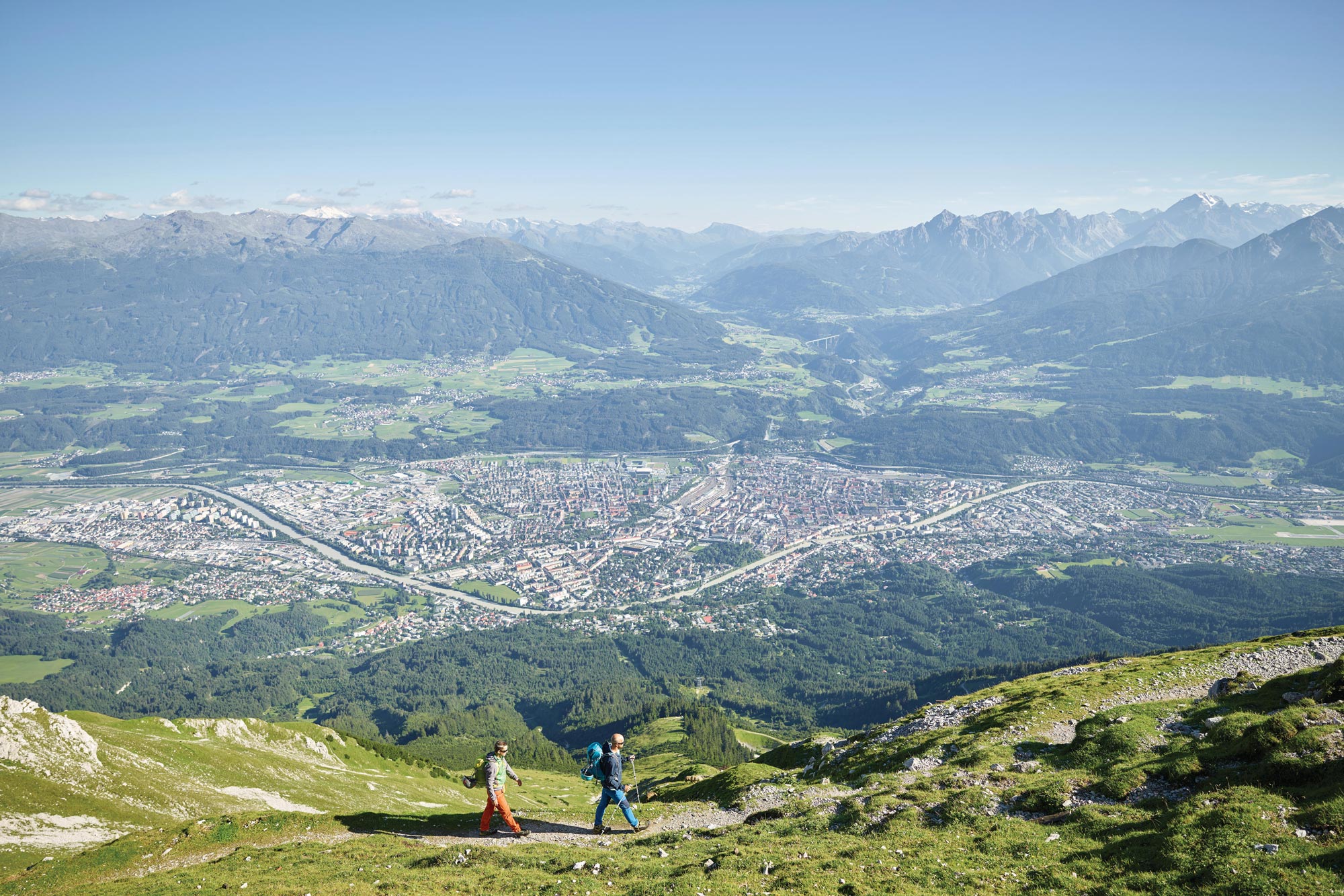 Due escursionisti su un sentiero verde di montagna con vista sulla città e sulle montagne.