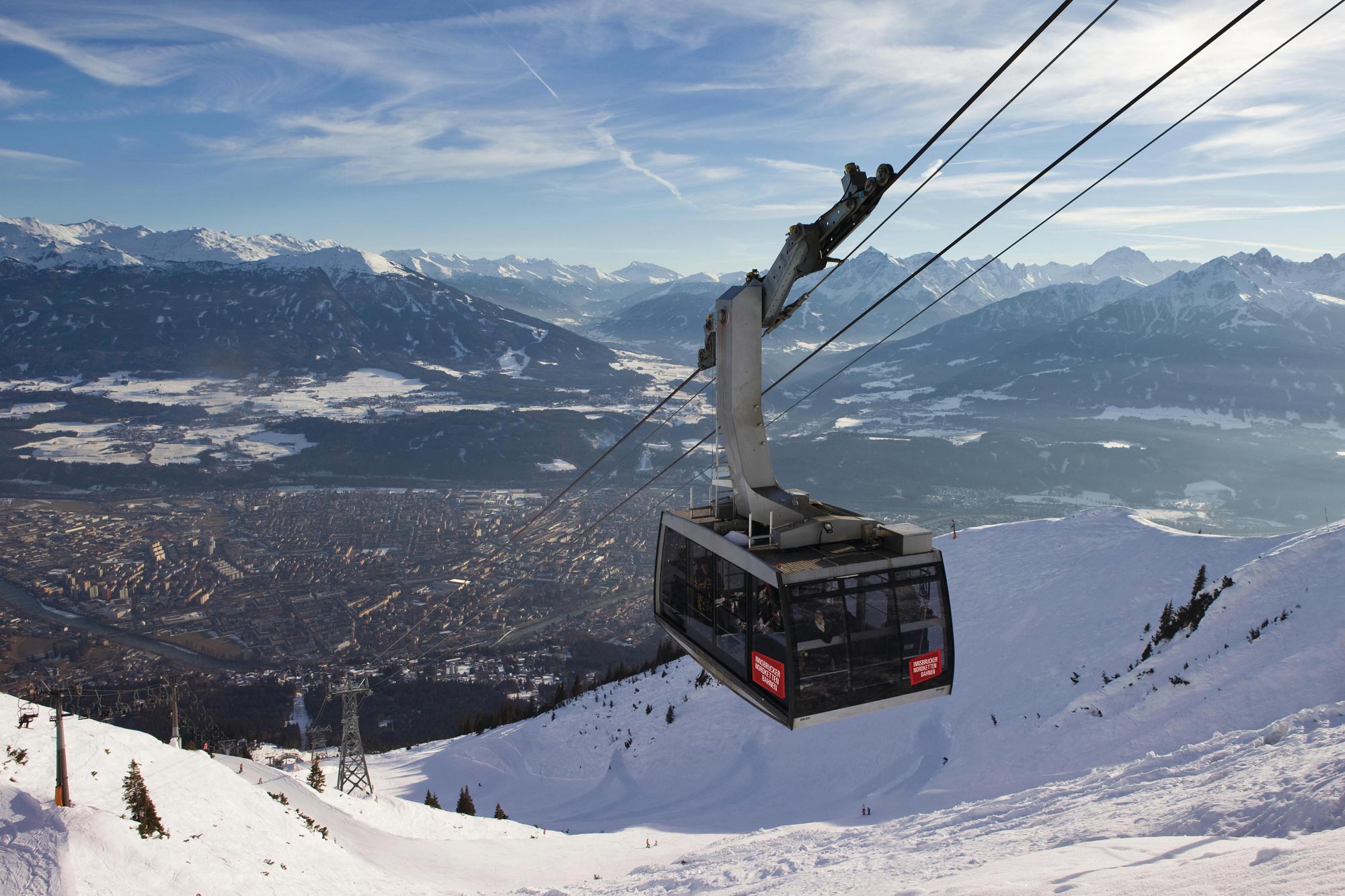 Cable car hovers over a snow-covered mountain landscape with a city panorama in the background.